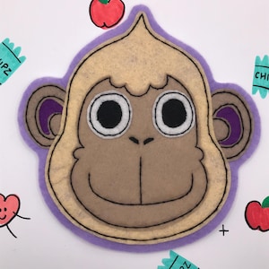 May include: A felt monkey face with a light brown face, large white eyes, and a purple outline. The monkey is surrounded by drawings of red apples, hearts, and teal-colored chip bags with the word "CHIPZ".