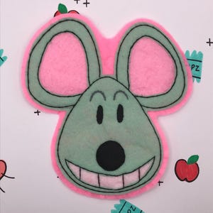 May include: A whimsical felt mouse face with a pink outline and ears, a green face, and a large, smiling mouth. The background features scattered black plus signs, stars, and small apple and candy illustrations.