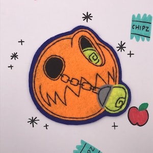 May include: Orange felt skull with a blue border, black details, and a chain. The skull has a green spiral in the eye and mouth. Two red apples and two teal "CHIPZ" packets are scattered around the skull.