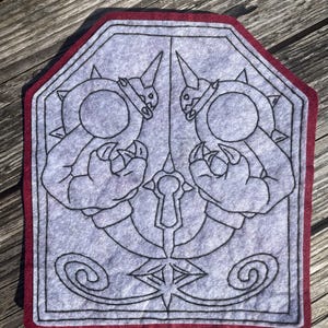 May include: Gray felt patch with a red border, featuring a black line drawing of two figures holding a keyhole. The figures have circular bodies, spiky details, and pointed horns. The design is symmetrical.