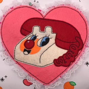 May include: A pink felt heart-shaped coaster featuring a cartoon telephone design. The telephone has a white body, red receiver, and blue eyes. The coaster is trimmed with white lace.