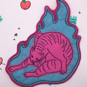 May include: A pink felt cat, with black outlines, is positioned within a blue flame-shaped border with a magenta edge. The background is white with doodles of apples, hearts, and crosses. The word "HIPZ" is visible.