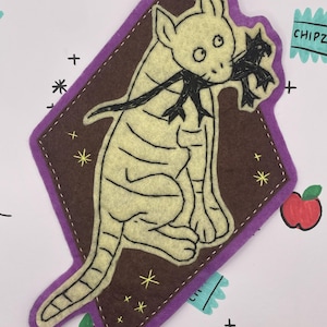 May include: A felt cat patch in a diamond shape. The cat is light yellow with black details, holding a black object in its mouth. The patch is on a brown background with a purple border. The background has drawn apples, stars, and the word "CHIPZ".