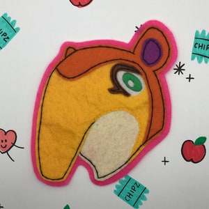 May include: A felt applique of a cartoon animal with orange, yellow, and white fur. The animal has a long snout, a green eye, and a purple ear. The applique is surrounded by a pink felt border.