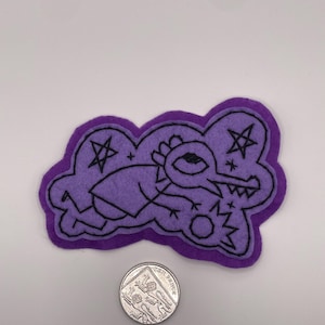 May include: A purple felt patch featuring black embroidered line art of a creature with a long snout and sharp teeth, surrounded by stars and a sun. A silver ten pence coin is placed below the patch for scale, showing its relative size.