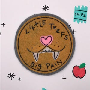 May include: A circular felt patch featuring a cartoon lion face. The patch is tan with a grey border, a pink heart-shaped nose, and the text "LITTLE TEETH BIG PAIN". The image also includes drawn apples, "CHIPZ" labels, asterisks, and plus signs.