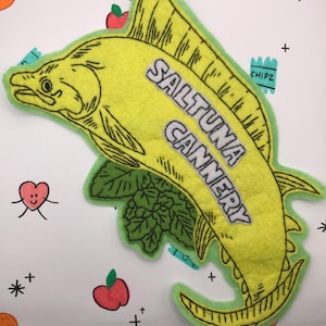May include: A yellow felt fish-shaped patch with the text "SALTUNA CANNERY" in white and black. The patch has a green border and is decorated with black outlines. The background is white with cartoon drawings of hearts, apples, and other shapes.