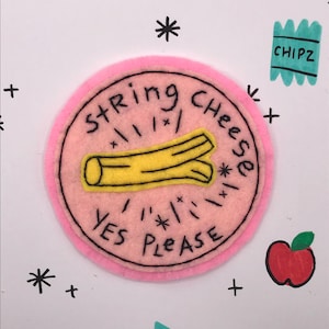 May include: A pink felt patch with the words "String Cheese YES PLEASE" around a yellow string cheese illustration. The background is white with drawn stars, plus signs, and two red apple illustrations.