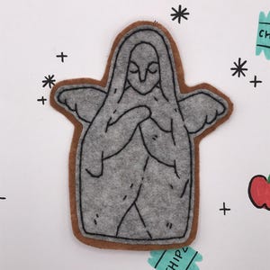 May include: A gray felt figure with a brown border, resembling a praying angel, is centered on a white background. The figure has folded hands and small wings. Black lines define the details. The background features red apple drawings, black crosses, asterisks, and a teal "CHIP" label.