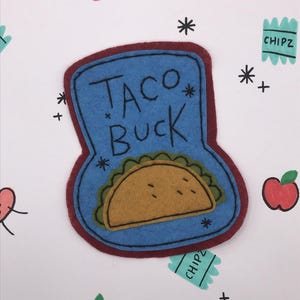 May include: A felt patch with the words "TACO BUCK" in black lettering, featuring a yellow taco with green lettuce. The patch is blue with a maroon border, surrounded by drawn apples, asterisks, and "CHIPZ" labels.