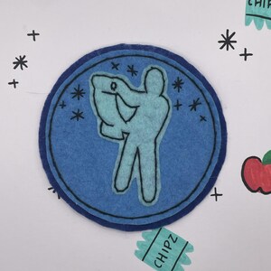 May include: A blue felt patch with a light blue silhouette of a person holding a bag, surrounded by stars. The patch is on a white background with additional star and plus symbols, and a "CHIPZ" label.