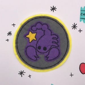 以下が含まれることがあります： 紫色のサソリと黄色の星が描かれたフェルトパッチ。灰色の円に黄色い縁取りが施されています。パッチは黒いアスタリスク、プラス記号、リンゴとスナックパッケージのイラストで装飾されています。