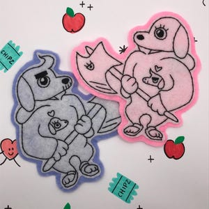 May include: Two felt patches featuring cartoon dogs holding axes. The dog on the left is light blue with a gray outline and the dog on the right is pink with a white outline. Both dogs have black eyes and a pink heart on their chest. The patches are on a white background with red apples, a green orange, and the word "CHIPZ" written on two green tickets.