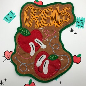 May include: A felt patch featuring the word "FRIENDS" in orange lettering on a brown background. Two red apples with white eyes and green leaves are stitched onto the patch. The patch is bordered in green and has a blue and white stitched design.