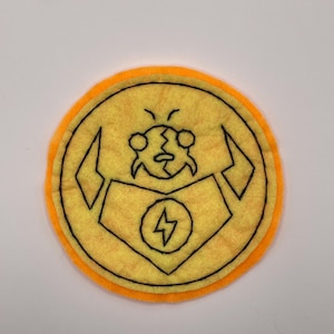 May include: A circular felt patch featuring a yellow centre with a black embroidered outline of a stylised character head and a chest plate displaying a lightning bolt symbol. The patch has a bright orange felt border around the outer edge.
