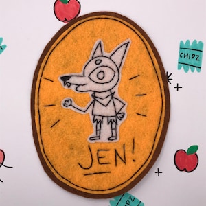 May include: A handmade felt patch featuring a cartoon wolf-like character with one large eye, outlined in black. The patch is yellow with a brown border and the name "JEN!" written below. Scattered around are drawn apples and "CHIPZ" packets.