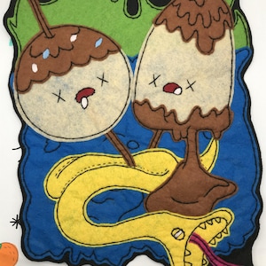 May include: A colourful felt artwork depicting two cartoon desserts with cross-shaped eyes and open mouths, and a yellow snake with a pink tongue. The desserts are brown and white, set against a blue background with green and black accents.