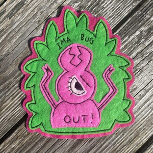 May include: A felt patch with a pink bug-like creature and the text "IMA BUG OUT!". The bug has a cracked head, one eye, and outstretched arms. The patch is outlined in black and set against a green leafy background with a pink border.
