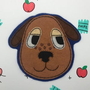 May include: A brown felt dog face with a blue felt border. The dog has white eyes, a black nose, and a brown mouth. The image is surrounded by hand-drawn red apples and blue and green "CHIPZ" labels.