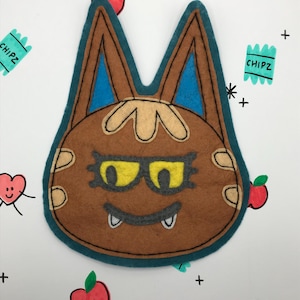 May include: A brown felt animal character with blue ears, yellow glasses, and a toothy grin. The character is surrounded by hand-drawn doodles of hearts, apples, and plus signs.