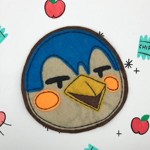 May include: A felt appliqué of a cartoon penguin with a blue head, brown eyes, and a yellow beak. The penguin is smiling and has orange cheeks. The appliqué is surrounded by a brown felt border.