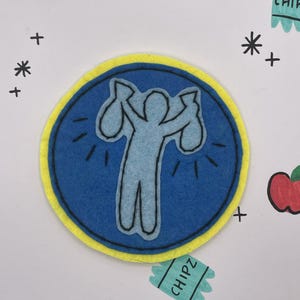 May include: A blue felt patch with a yellow border features a light blue figure holding two bags. The patch is surrounded by drawn stars, plus signs, and two small green rectangles with the word "CHIPZ".