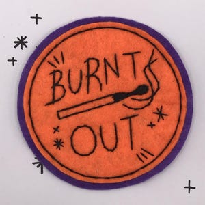 May include: An orange felt patch with a purple border. The patch features the words "BURNT OUT" in black lettering, along with a drawing of a burnt match. Decorative stars and plus signs surround the patch.