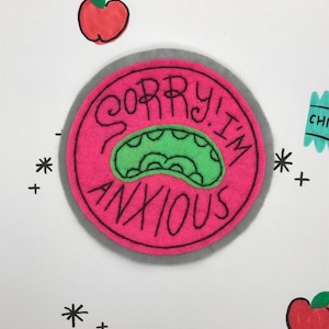 May include: Pink felt patch with a gray border and the text "Sorry I'm Anxious" in black embroidery. A green cartoon mouth with teeth is inside the text.