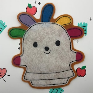 May include: A felt appliqué of a friendly ghost with rainbow hair. The ghost is smiling and has two small arms. The ghost is surrounded by a brown felt border.