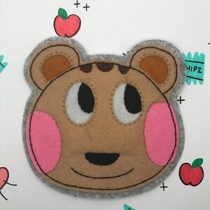 May include: A brown felt animal face with pink cheeks and a black nose. The face is surrounded by a gray felt border. The animal is wearing a gray felt hat with brown felt ears. The background is white with red apples and the text "CHIPZ" in green.