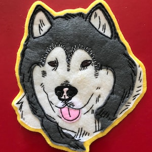 May include: A felt appliqué of a husky dog's face with black and gray fur, a pink tongue, and a yellow border.
