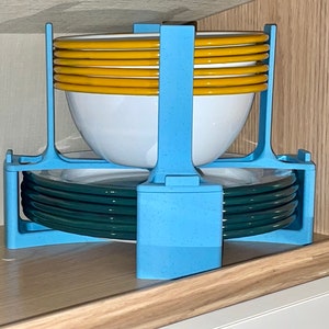 Op de afbeelding: Een blauwe plastic serviesrek met een stapel gele en witte kommen en een stapel donkergroene borden.