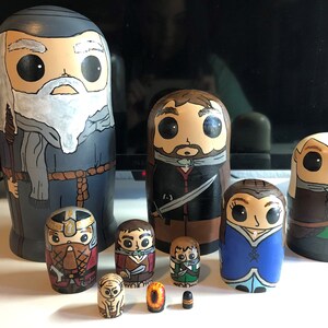 May include: A collection of hand-painted wooden nesting dolls, each depicting a character with large eyes and stylized features. The dolls vary in size, with the largest being approximately 8 inches tall, and feature a variety of colors and designs.