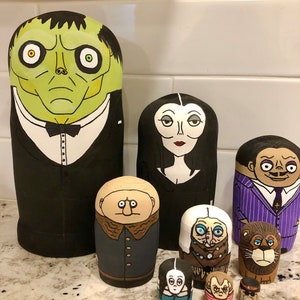 May include: A collection of hand-painted wooden nesting dolls featuring characters from The Addams Family. The dolls are painted in vibrant colors and detailed designs, including Gomez, Morticia, and Lurch. The largest doll is approximately 10 inches tall.