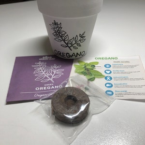 May include: A white ceramic pot with a black drawing of oregano leaves and the word "OREGANO" on the side. A purple packet with the words "URBAN LEAF GREEK OREGANO" and a small brown seed disc are also in the image. A white card with the word "OREGANO" and a list of health benefits is in the background.