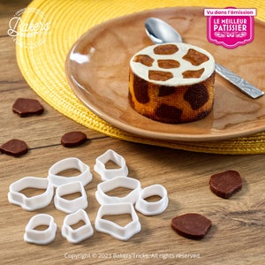 Peut inclure: Des emportes-pièces en plastique blanc de différentes formes, y compris des cercles, des ovales et des diamants. Les emportes-pièces sont disposés sur une surface en bois. Il y a aussi plusieurs morceaux de chocolat brun sur la surface. Une assiette brune avec un gâteau à motif de girafe blanc et brun est en arrière-plan.