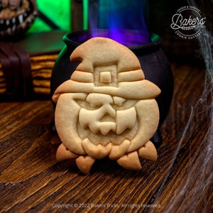May include: A witch cookie cutter in the shape of a witch's face with a hat. The cookie cutter is made of metal and has a shiny finish. The cookie cutter is sitting on a wooden table next to a cauldron.