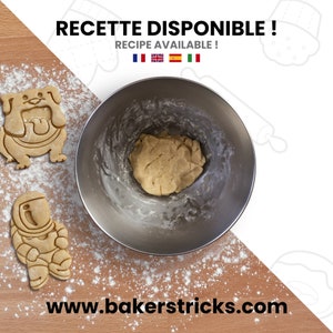 May include: A stainless steel mixing bowl with cookie dough inside. The bowl is on a wooden surface covered in white flour. There are two cookie cutters on the surface, one shaped like a bulldog and the other shaped like an astronaut. The text "RECETTE DISPONIBLE! RECIPE AVAILABLE!" is at the top of the image.