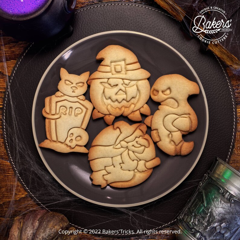 May include: Four Halloween-themed cookie cutters on a black plate. The cutters are shaped like a pumpkin, a ghost, a witch, and a cat with a tombstone. The cutters are made of metal and have a shiny finish.
