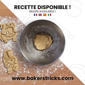 May include: A stainless steel mixing bowl with cookie dough inside. The bowl is on a wooden surface covered in white flour. There are two cookie cutters on the surface, one shaped like a bulldog and the other shaped like an astronaut. The text "RECETTE DISPONIBLE! RECIPE AVAILABLE!" is at the top of the image.