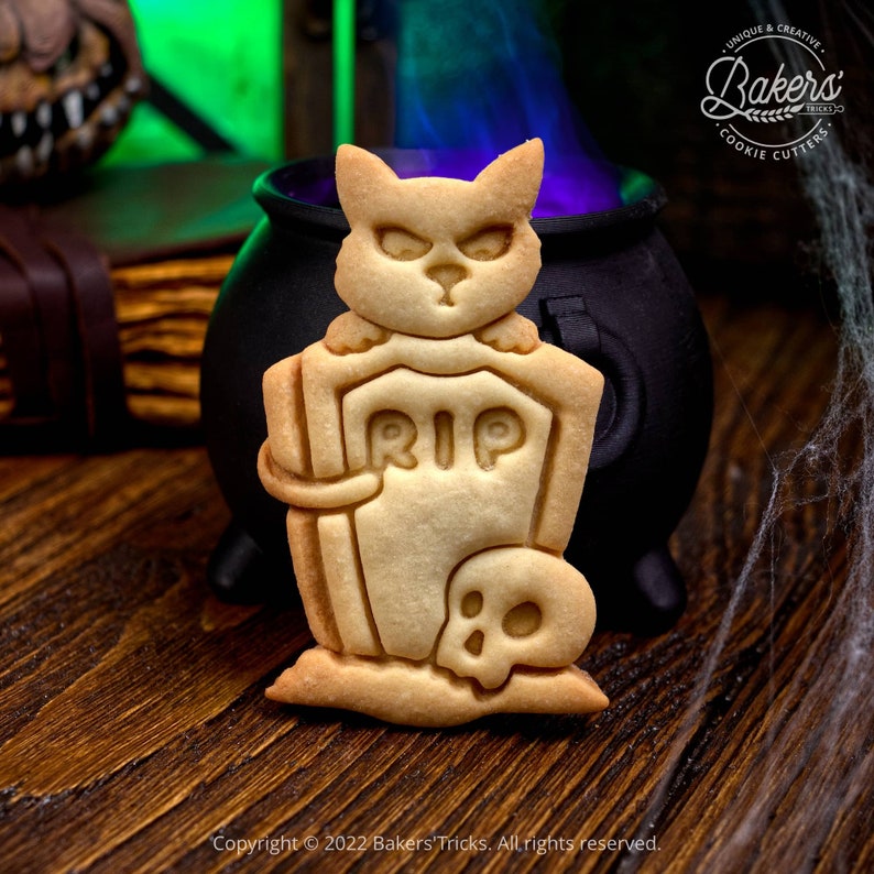 May include: A Halloween cookie cutter in the shape of a cat sitting on a tombstone with a skull at the base. The cookie cutter is made of metal and has a gold finish. The cookie cutter is sitting on a wooden surface.