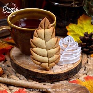 A baked leaf-shaped cookie and a white plastic leaf cookie cutter rest on a wood slice next to a mug of tea and autumn decor.