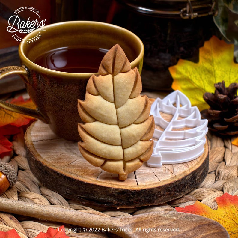 A baked leaf-shaped cookie and a white plastic leaf cookie cutter rest on a wood slice next to a mug of tea and autumn decor.