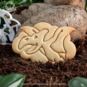 Peut inclure: Un biscuit brun clair avec un motif de dinosaure. Le biscuit est posé sur un lit de terre brune avec des plantes vertes en arrière-plan.