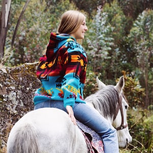 May include: A person wearing a turquoise, red, and orange patterned hoodie and blue jeans, riding a gray horse. The horse has a white mane and a brown bridle. The background features green trees and a rocky hillside.