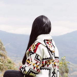May include: A person wearing a white hooded sweater with a geometric pattern in black, red and yellow. The person is sitting on a grassy hillside and looking out at the view.