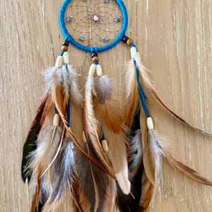 May include: A handcrafted dreamcatcher with a turquoise blue hoop and a web of brown string. The dreamcatcher features brown and tan feathers, wooden beads, and small decorative stones. The feathers are attached with blue string.
