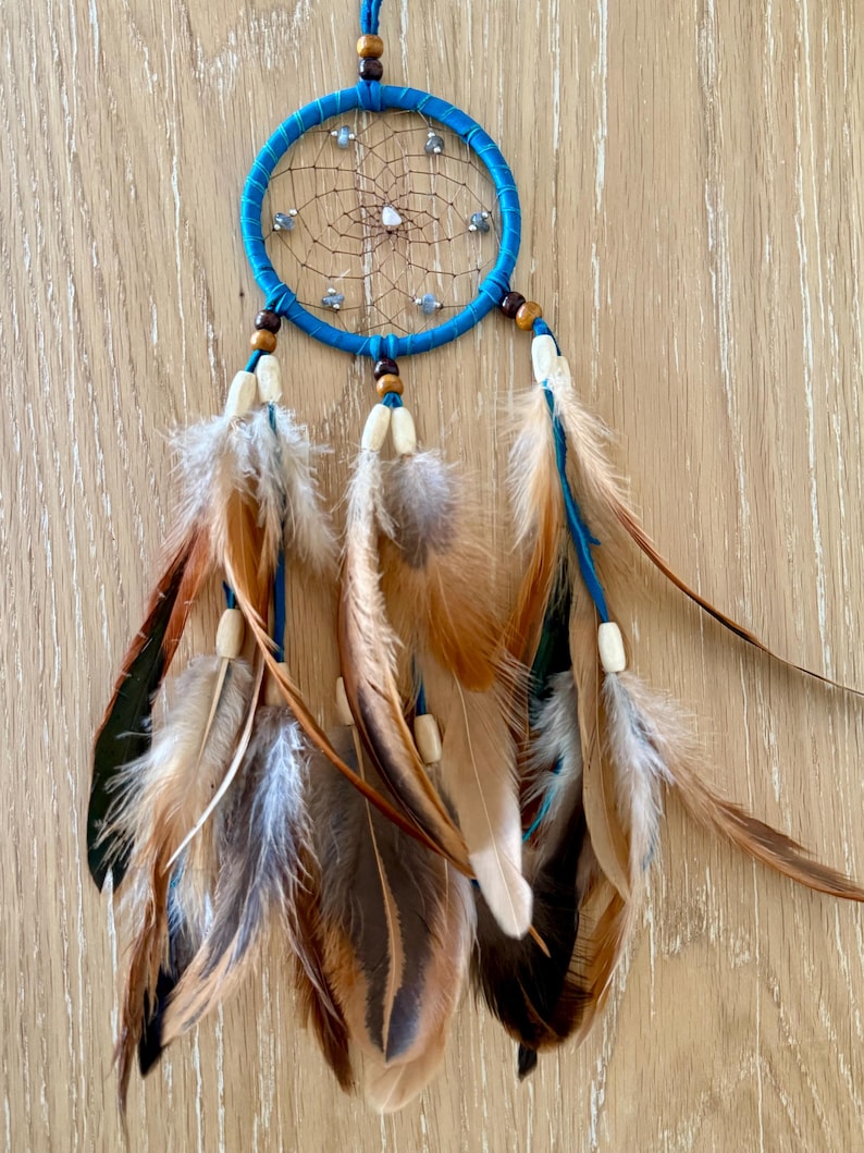 May include: A handcrafted dreamcatcher with a turquoise blue hoop and a web of brown string. The dreamcatcher features brown and tan feathers, wooden beads, and small decorative stones. The feathers are attached with blue string.