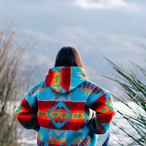 Könnte beinhalten: Eine Person in einem blauen, roten, orangen und gelben gemusterten Kapuzenpullover sitzt mit dem Rücken zur Kamera auf einem Felsen. Die Person blickt auf ein Gewässer und Berge in der Ferne.