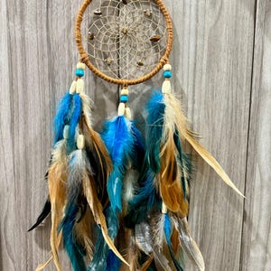 May include: A handcrafted dreamcatcher with a circular, woven web design. The dreamcatcher features a brown wooden hoop, adorned with small stones and hanging feathers in shades of blue, teal, and tan. Decorative beads are also included.